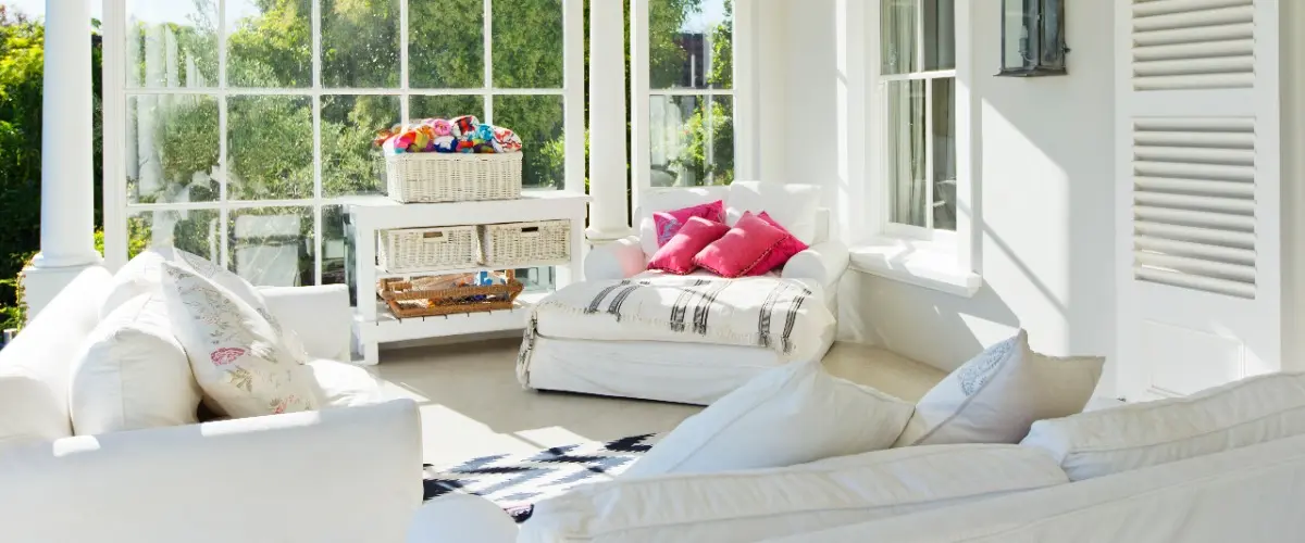 A bright white sunroom featuring white furniture and pillows, creating a serene and airy atmosphere.
