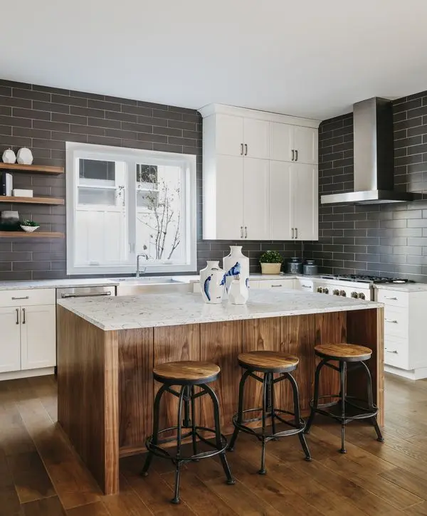 A modern kitchen featuring dark grey subway tile walls, white upper cabinets, and a natural wood island with white quartz countertops and industrial bar stools.