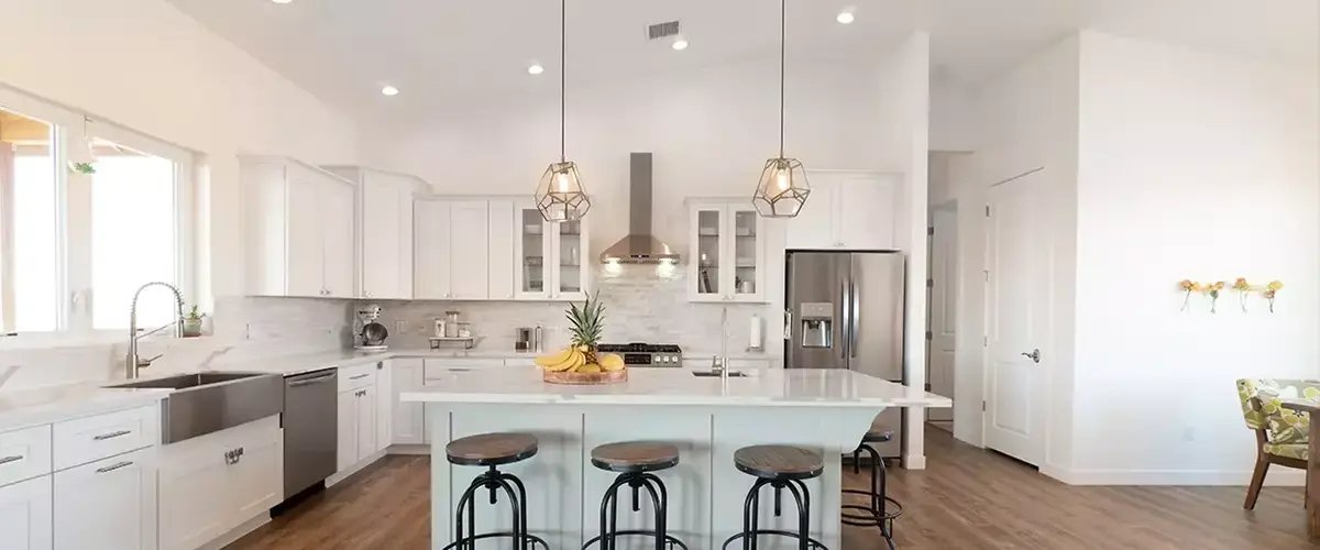 An airy, open-concept white kitchen with high ceilings, a large light blue island with seating, and modern geometric pendant lights hanging above.