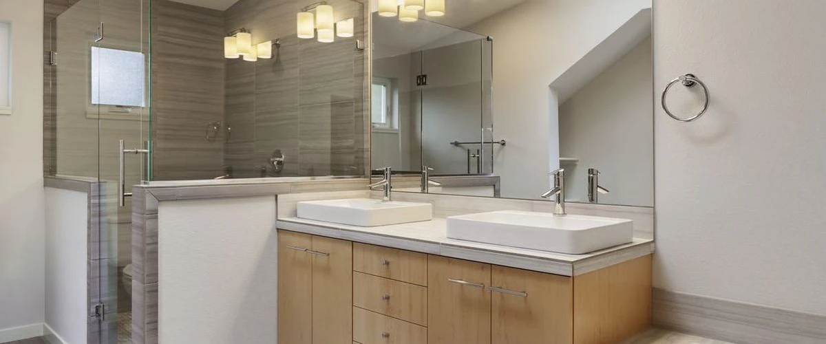 A contemporary bathroom interior showcasing a light wood double vanity with two white vessel sinks, modern chrome faucets, and a large wall-to-wall mirror reflecting a glass shower.