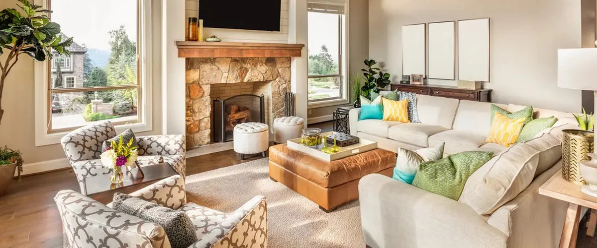 A sunlit contemporary living space showing a large stone fireplace, a brown leather ottoman coffee table, a cream sectional sofa with colorful pillows, and large windows overlooking greenery.