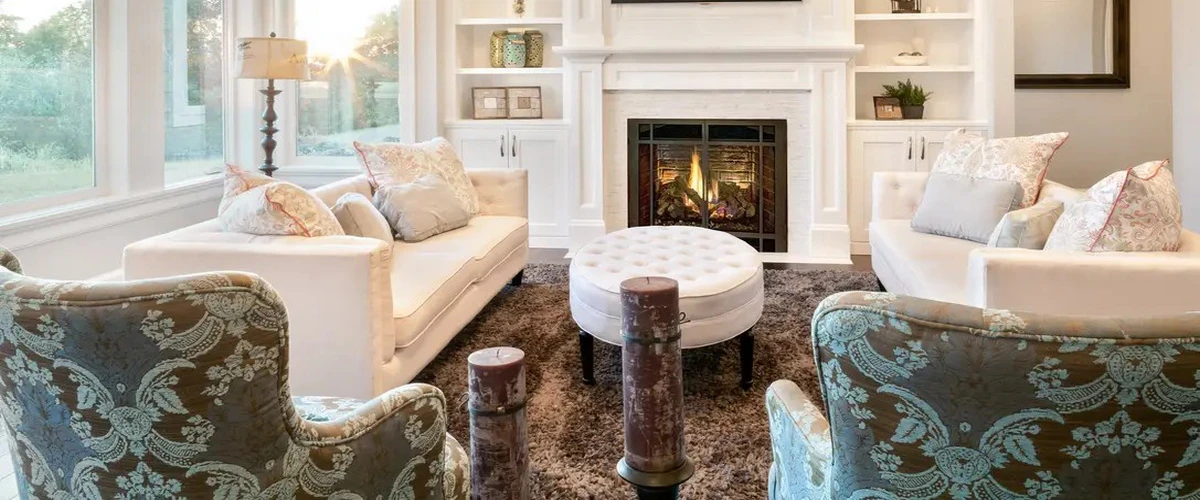 A bright, elegant living room featuring two white sofas, patterned teal armchairs, a central tufted ottoman, and a glowing fireplace set against white built-in shelving.