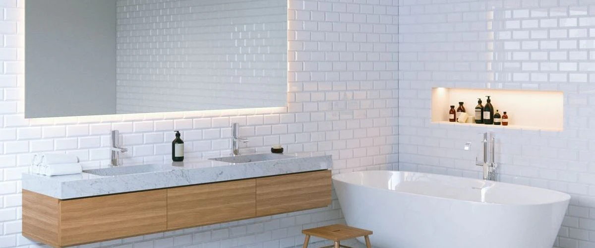 Minimalist bathroom design showcasing white subway tile walls, a floating light wood vanity with a marble double sink, and a sleek white freestanding bathtub next to a recessed wall niche with toiletries.