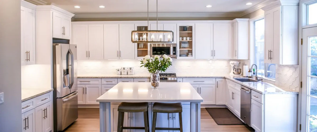 A bright, modern kitchen featuring white shaker cabinets, a large central island with grey barstools, stainless steel refrigerator, and elegant hanging pendant lighting.