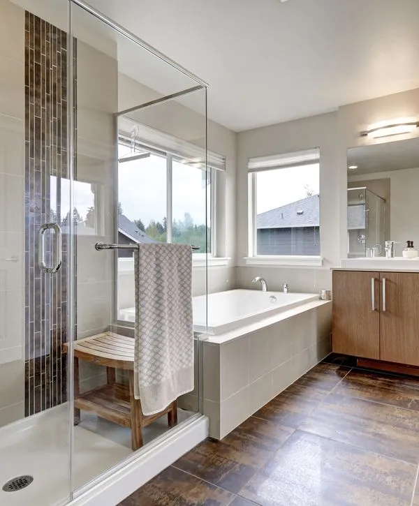 A luxury primary bathroom remodeling in La Vista, NE featuring a glass-enclosed walk-in shower with a wooden bench and a large white soaking tub under a window.