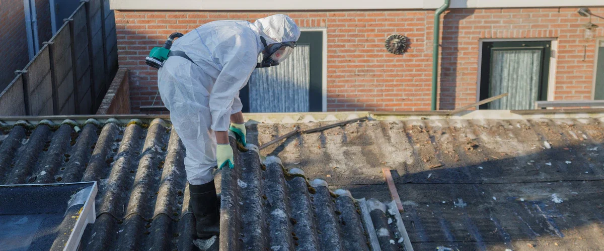 A worker in a white hazmat suit and full-face respirator mask leans over to inspect old, weathered asbestos roofing panels on a residential building.