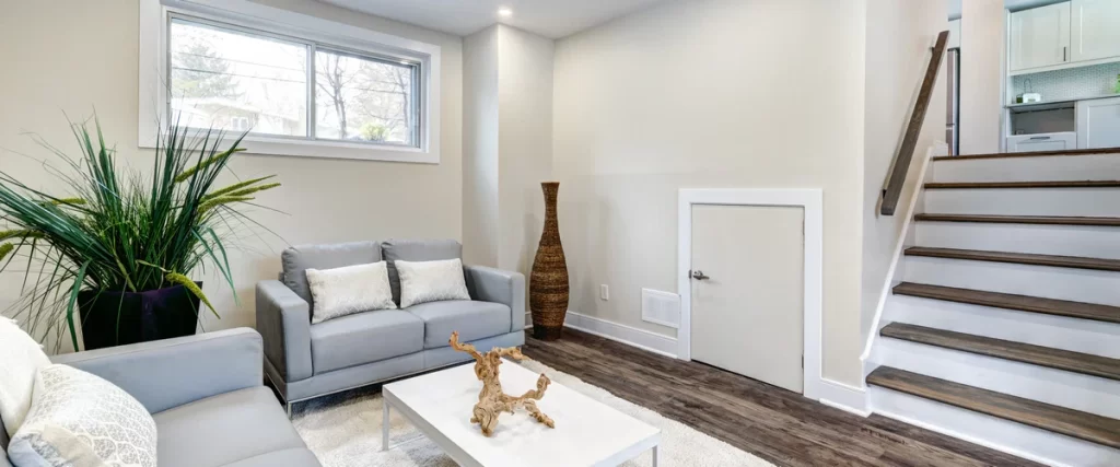 A bright, completed basement finishing project showcasing a modern living area with grey sofas, a white coffee table, and a wooden staircase.