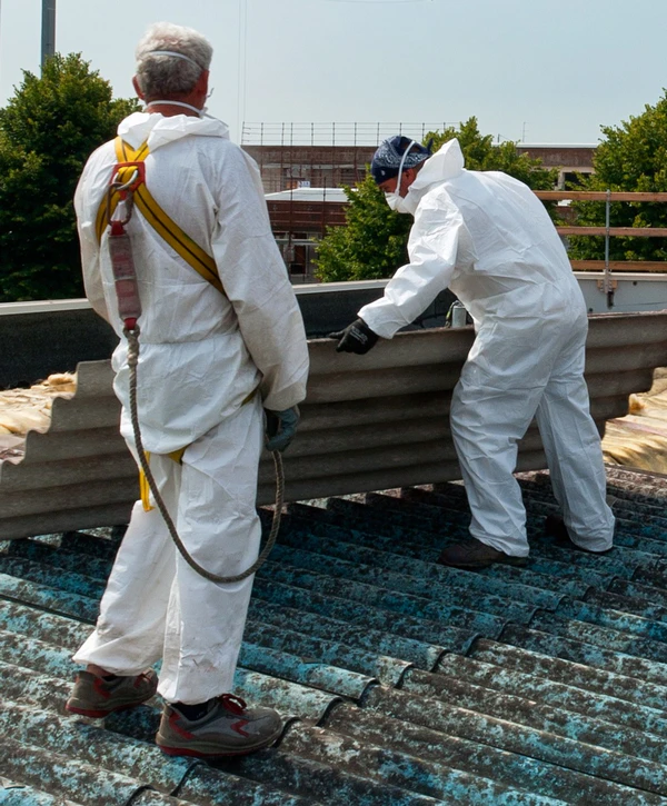 Two workers wearing white protective hazmat suits and a safety harness carefully lifting a grey corrugated sheet during an asbestos removal project.