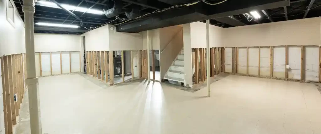 A wide interior perspective of a clean, unfinished basement with exposed wooden stud walls, concrete floor, visible furnace, water heater, a central staircase, and overhead wiring, ready for a finishing contractor.
